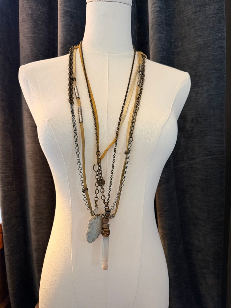 A multi-layered bohemian-style necklace displayed on a white mannequin torso. The necklace features antique bronze and gold chains, a light brown woven cord, a silver-toned cylindrical charm, and a large natural gray stone pendant with a clear quartz crystal drop.