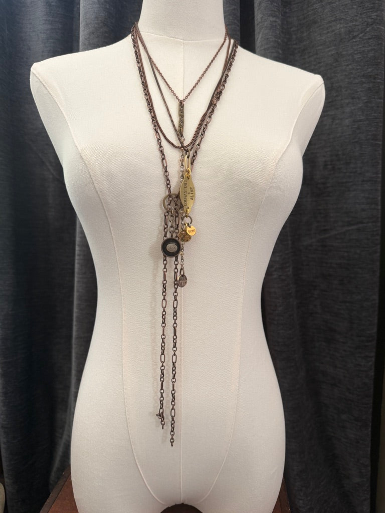 A long layered necklace with mixed metallic finishes displayed on a white dress form mannequin against a dark gray curtain background.