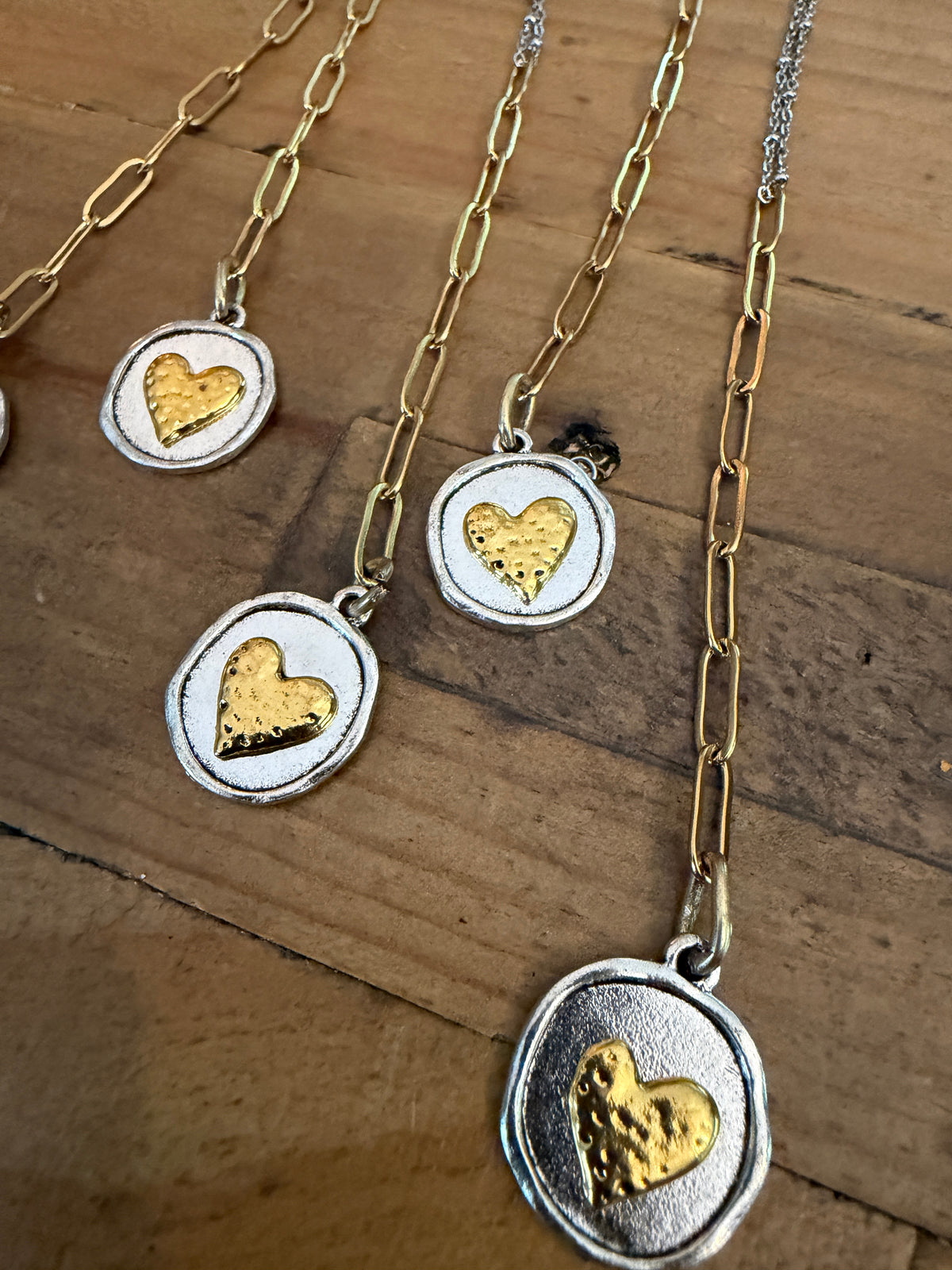 Three silver and gold necklaces with oval pendants featuring textured gold hearts, displayed on a rustic wooden surface.