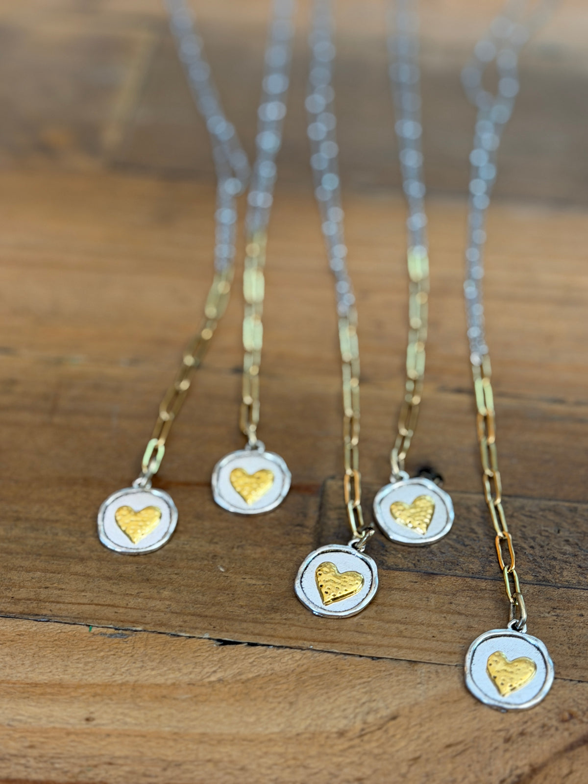 Four silver and gold necklaces with round pendants featuring textured gold hearts, arranged on a rustic wooden surface.