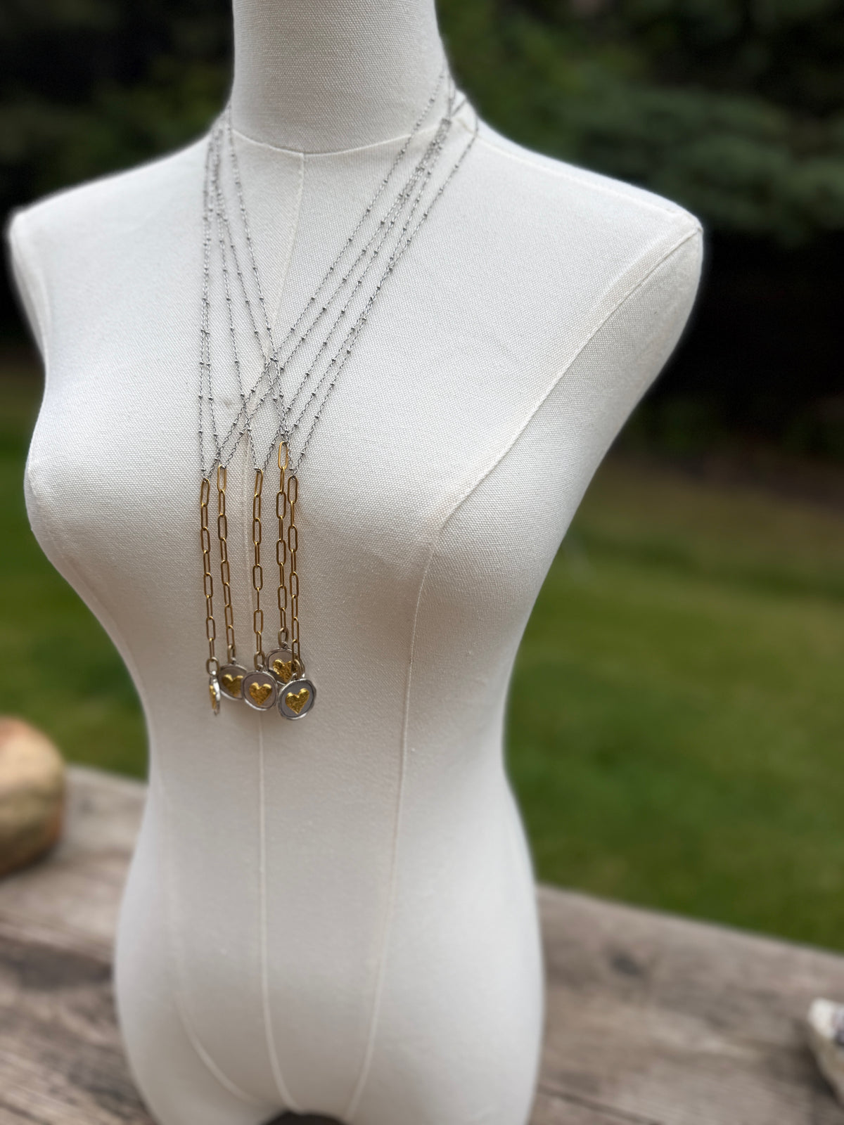 A layered necklace displayed on a white dress form mannequin outdoors. The necklace features multiple silver-toned chains, gold-toned chain elements, and three dangling charms, including a round amber-colored bead and two smaller oval charms.