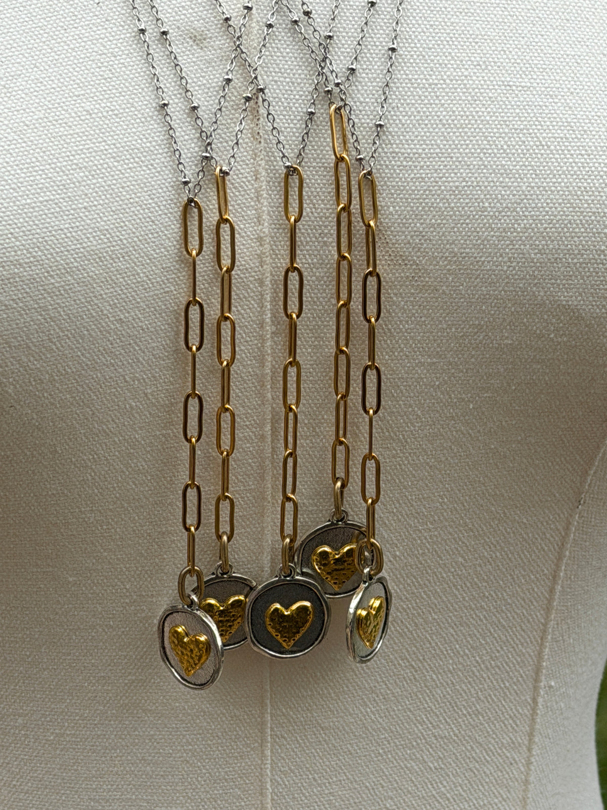 Three gold and silver paperclip chain necklaces with oval heart pendants displayed on a cream fabric mannequin torso.