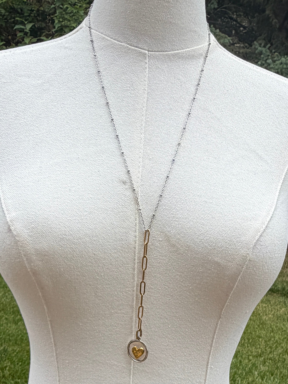 A long silver chain necklace with a small gold heart pendant displayed on a cream fabric mannequin torso outdoors, with grass and trees in the background.