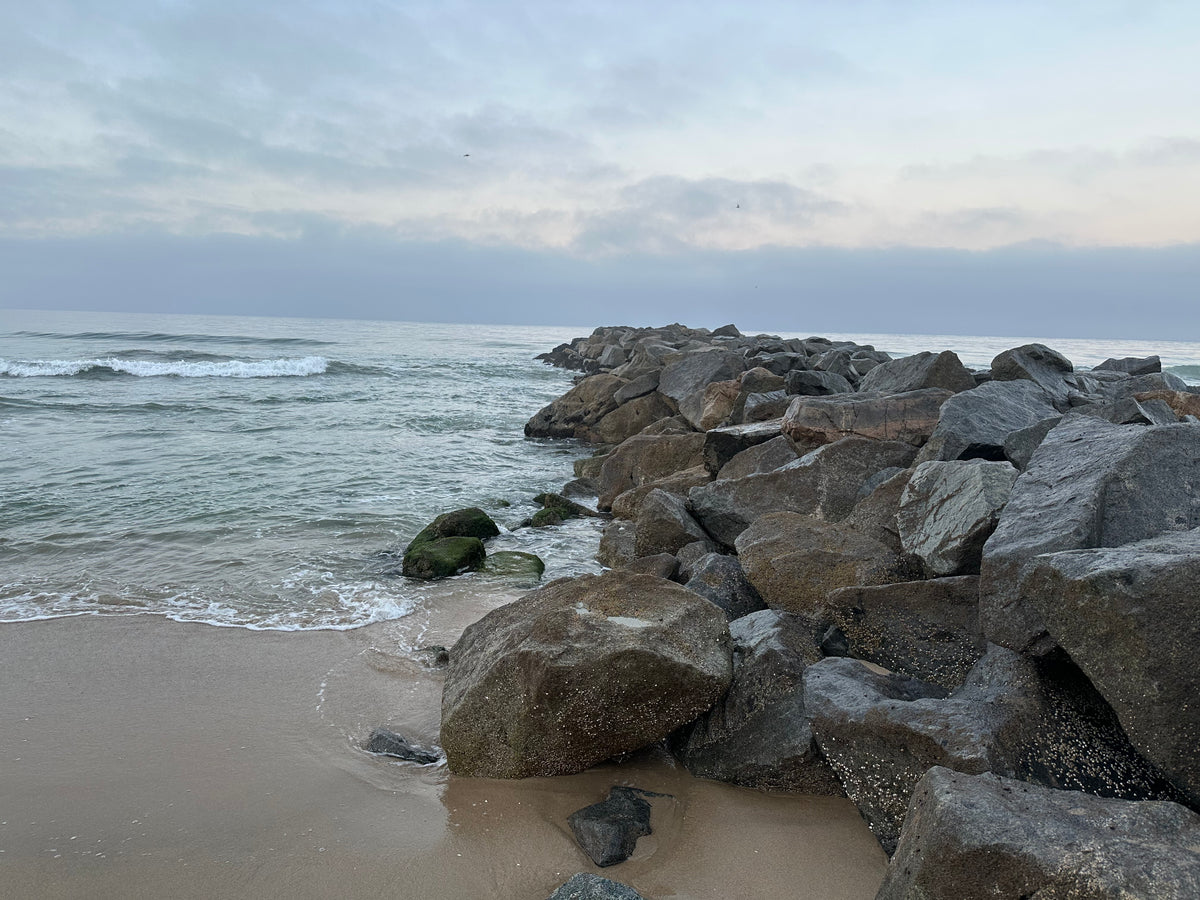 A coastal scene with large dark rocks forming a breakwater beside a sandy beach and calm ocean water under a cloudy sky.