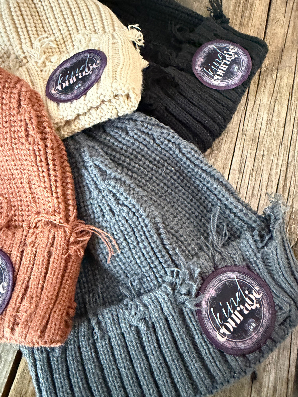 Four knitted hats in black, gray, beige, and brown with branded buttons on a wooden surface.