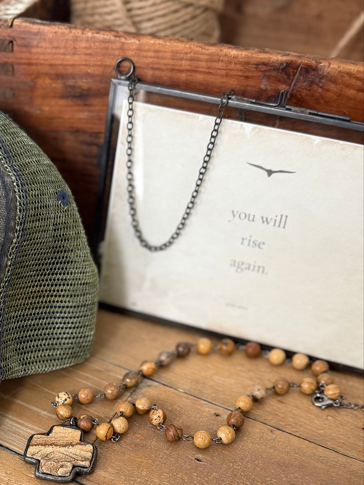 Green cap with 'True North' patch, wooden cross necklace, and rustic wooden box on a wooden surface.