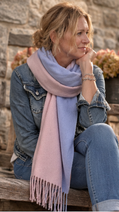A woman sitting outdoors on a wooden bench wearing a soft pastel pink and light blue scarf, a denim jacket, and blue jeans, with a stone wall and flowers in the background.