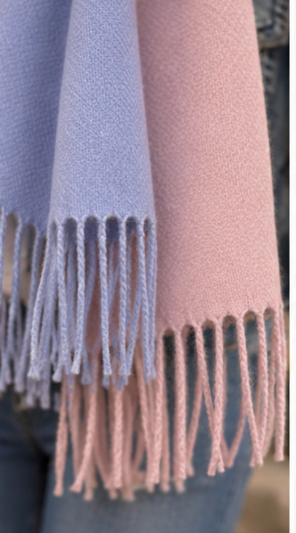 A close-up of a soft woven scarf with fringed ends, shown draped over blue jeans. The scarf appears in two pastel colors: a light lavender-blue on the left and a dusty pink on the right.