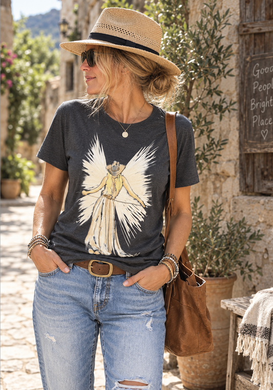 A woman wearing a dark charcoal graphic t-shirt with a cowboy angel illustration, paired with light-wash distressed jeans, a straw hat, and a brown leather shoulder bag, standing in a rustic outdoor setting.