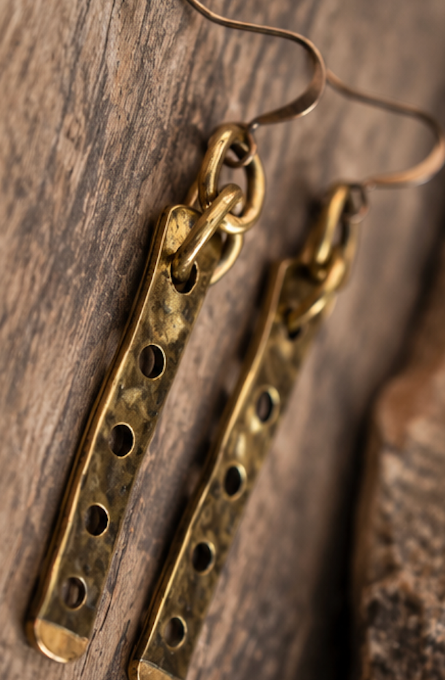 A pair of handmade brass dangle earrings with elongated rectangular bars featuring five circular cutouts, resting on a rustic wooden surface.